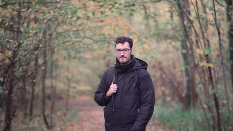Engaging-bearded-European-man-with-sharp-glasses,-standing-in-a-mixed-autumn-forest,-enthusiastically-pointing-and-beckoning-viewers-closer,-inviting-them-to-join-a-nature-exploration-journey