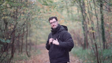 Engaging-bearded-European-man-with-sharp-glasses,-standing-in-a-mixed-autumn-forest,-enthusiastically-pointing-and-beckoning-viewers-closer,-inviting-them-to-join-a-nature-exploration-journey