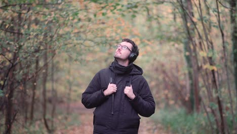 Young-European-man-with-headphones-in-a-vibrant-fall-mixed-forest,-observing-colorful-autumn-leaves