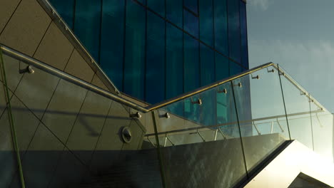Modernes-Gebäude-Mit-Blau-Getönten-Fenstern-Und-Elegantem-Glasgeländer-Unter-Einem-Sonnenbeschienenen-Himmel