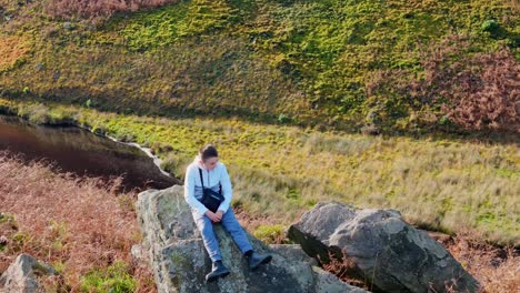 A-young-lad-takes-a-moment-on-a-large-rock-outcrop,-lost-in-thought,-while-aerial-footage-revolves-around-the-moorland