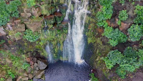 Yorkshire-Moors-striking-waterfall,-aerial-view-captures-water-flowing-over-large-rocks-into-a-deep-blue-pool,-hikers-nearby