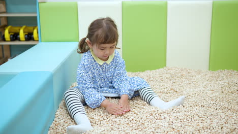 Toddler-girl-play-in-dry-pool,-rake-soft-Hinoki-wood-cubes-making-heap-or-pile-at-children's-entertainment-center,-tactile-sensors-development