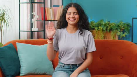 Indian-woman-smiling-friendly-at-camera,-waving-hands-gesturing-hello,-hi,-greeting-at-home-on-sofa