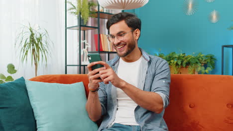 Indisch-arabischer-Junger-Mann,-Der-Auf-Dem-Sofa-Sitzt,-Nutzt-Sein-Smartphone-Und-Teilt-Nachrichten-In-Einer-Social-Media-Anwendung