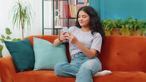 Indisch-arabische-Junge-Frau,-Die-Auf-Dem-Sofa-Sitzt,-Nutzt-Ihr-Smartphone-Und-Teilt-Nachrichten-In-Einer-Social-Media-Anwendung