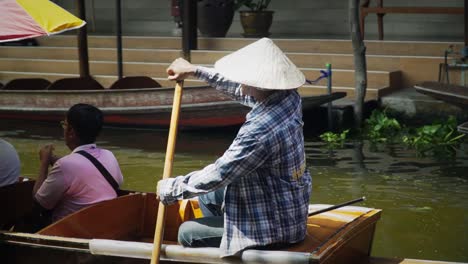 Helmsman-driving-the-passenger-boat-using-paddle-through-the-canal-in-Bangkok,-Thailand-handheld-panning