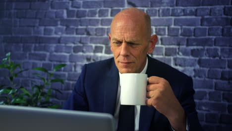 Happy-mature-bald-business-man-taking-a-break-at-work-with-a-coffee-sitting-in-front-of-a-laptop-in-an-office