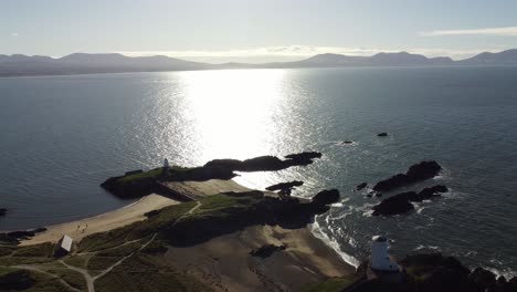Aerial-reversing-view-over-Ynys-Llanddwyn-Welsh-island-with-shimmering-ocean-and-misty-Snowdonia-mountain-range-across-the-sunrise-skyline