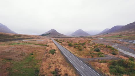 Aerial:-Reveal-of-silver-SUV-traveling-along-the-Iceland-Ring-Road-which-is-a-scenic-highway-through-a-picturesque-remote-fjord-area-leading-to-twin-peaks-with-fog-and-haze-in-the-distance