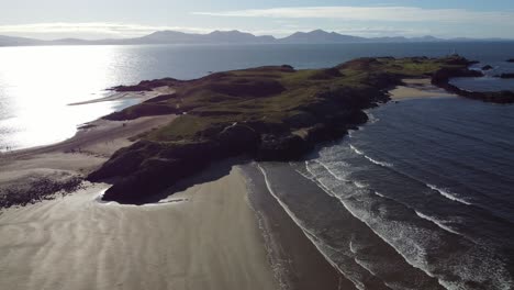 Wide-aerial-view-circling-Ynys-Llanddwyn-Welsh-island-with-shimmering-ocean-and-misty-Snowdonia-mountain-range-across-the-sunrise-skyline