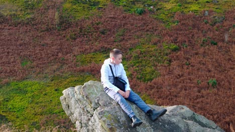 A-young-boy-sits-on-a-large-rock-outcrop,-contemplating-the-moorland-surroundings