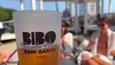Enjoying-a-nice-cold-beer-with-friends-in-Bibo-Beach-House-Tarifa-restaurant,-blurry-people-in-restaurant-of-Dani-Garcia-in-Spain,-fun-sunny-day,-4K-shot