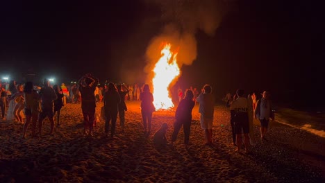 People-dancing-at-traditional-bonfire-summer-festival-at-the-beach-at-the-San-Juan-celebration-in-Marbella-Spain,-enjoying-a-fun-party,-big-burning-fire-and-hot-flames-at-night,-4K-shot