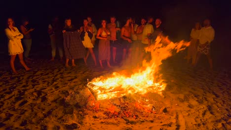 People-jumping-over-traditional-bonfire,-summer-festival-at-the-beach-at-the-San-Juan-celebration-in-Marbella-Spain,-enjoying-a-fun-party,-big-burning-fire-and-hot-flames-at-night,-4K-shot