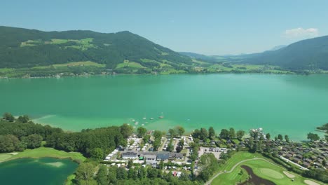 Lujosa-Orilla-Del-Lago:-El-Dron-Muestra-La-Tranquilidad-Del-Lago-Mondsee,-Abrazado-Por-La-Grandeza-De-Las-Montañas-Austriacas,-Ofreciendo-Una-Visión-De-Un-Campo-De-Golf.