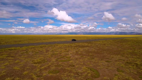 Drone-shot-flying-around-a-grey-car-driving-on-a-straight-road-through-the-Salinas-Grandes-salt-lake-on-the-border-of-Salta-and-Jujuy,-Argentina