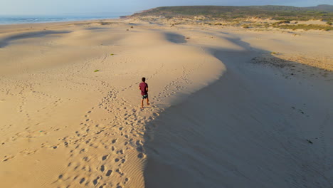Aerial-view-of-young-man-walking-on-dune-during-sunset-on-Bordeira-beach,-Portugal
