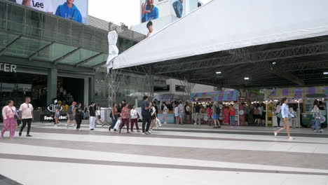 People-are-busy-walking-outside-of-Siam-Paragon-in-Bangkok-with-the-BTS-train-station-with-trains-leaving-and-stopping-for-passengers,-in-Thailand