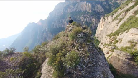 FPV-aerial-flying-circles-around-a-drone-pilot-perched-on-a-rocky-ledge-in-the-mountains
