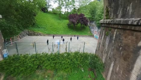 FPV-aerial-flying-under-an-aqueduct-and-circling-a-group-of-guys-playing-football-on-an-inner-city-court