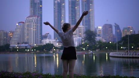 A-woman-raises-her-arms-in-celebration-in-front-of-a-city-across-a-lake