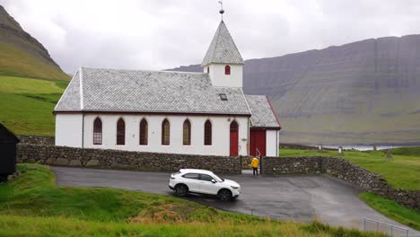 Mann-Betritt-Den-Hof-Der-Weißen-Vidareidi-Kirche-Auf-Einer-Wiese-In-Den-Bergen,-Färöer-Inseln