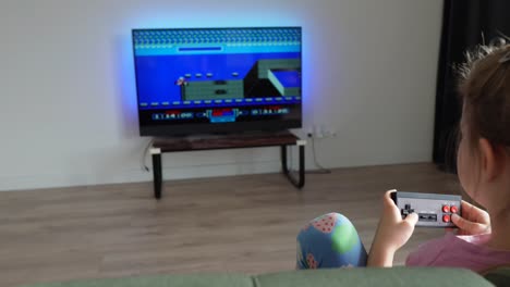 Toddler-girl-plays-with-vintage-gaming-console-during-the-evening,-moving-cinematic-shot,-TV-with-a-game-in-the-background