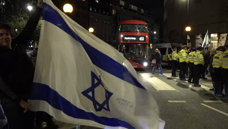 Ein-Mann-Hält-Eine-Große-Blau-weiße-Israelische-Flagge-In-Der-Hand,-Während-Ein-Verbindungsbeamter-Der-Stadtpolizei-Während-Einer-Protestaktion-Einen-Roten-Londoner-Bus-Auf-Der-Straße-Vorbeischwenkt