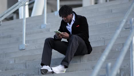 Junger-Afrikanischer-Geschäftsmann-Sitzt-Auf-Einer-Treppe-Und-Schreibt-Eine-SMS-Auf-Seinem-Handy