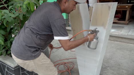 A-middle-aged-man-painting-in-white-color-a-wooden-surface-with-a-spray-gun-outside-a-carpentry-workshop