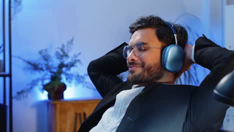 Happy-smiling-Indian-businessman-listening-music-on-headphones-taking-break-relaxing-at-office