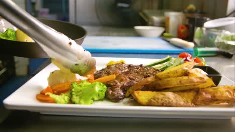 Chef-using-metal-tongs-to-place-grilled-vegetables-on-plate-with-steak-and-potatoes