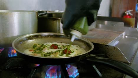 Chef-wearing-black-rubber-gloves-pours-green-mixture-into-cooking-pan-with-white-sauce-and-tomatoes
