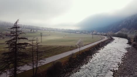 Moody-foggy-rural-aerial-scenery,-fields-with-farm-houses-and-a-road-with-cars,-flying-up-Ötztal,-Austria