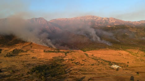 Aerial-footage-of-dramatic-wildfires-burning-in-the-mountains-on-the-outskirts-of-the-city-of-Vlorë,-Albania,-lit-by-an-orange-sunset
