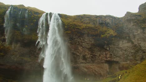 Stunning-tilt-down-of-Seljalandsfoss-waterfall-to-tourists-hiking-on-path-near-mountain