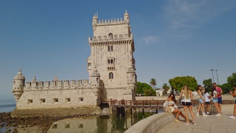 Turistas-Tomando-Fotografías-Con-La-Torre-De-Belem-Y-Su-Muelle-De-Madera-Al-Fondo,-Lisboa,-Portugal