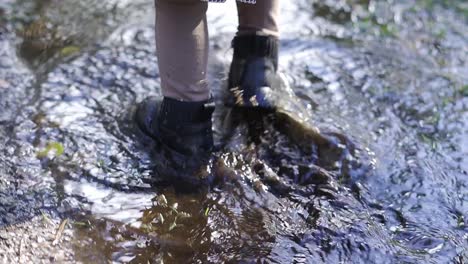 Close-up-of-toddler-walking-in-a-mud-puddle-with-her-black-winter-boots,-splashing-in-the-autumn-forest