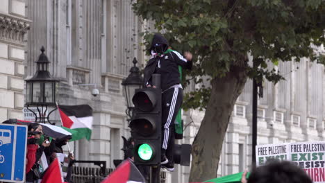 A-masked-protestor-sits-on-a-traffic-light,-joins-in-chants-and-points-to-the-sky,-as-others-wave-flags-and-placards-outside-Downing-Street-on-Whitehall-at-a-national-demonstration-for-Palestine