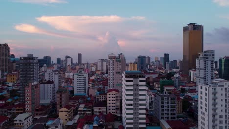 Drone-view-over-Phnom-Penh-city-skyline-in-the-evening,-aerial-drone-shot,-trucking-left,-roof-level,-with-distant-thunderstorm-cloud-in-the-distance-and-bright-vivid-colours-of-the-buildings