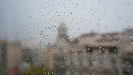 A-slow-motion-close-up-view-of-heavy-raindrops-seen-through-a-window,-with-an-urban-city-landscape-in-the-background