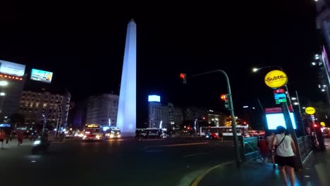 POV-Nachtansicht-Des-Obelisken-In-Der-Stadt-Buenos-Aires-Mit-Stau-Und-Menschen,-Die-Die-U-Bahn-Betreten