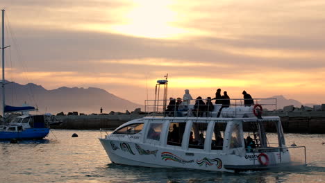 Hermanus-whale-watching-boat-full-of-tourists-cruising-out-of-harbor-at-sunrise