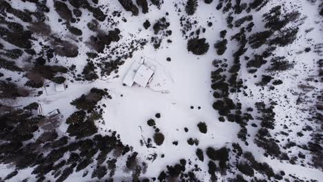 Aerial-top-down-winter-snow-covered-mountain-slope,-trees-glowing-and-snowy-hut-cabin-Lehnberghaus-in-Austria,-bird's-eye-view-flying-down