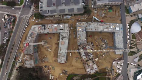 Top-down-drone-view-of-large-construction-site-with-cranes-moving-below