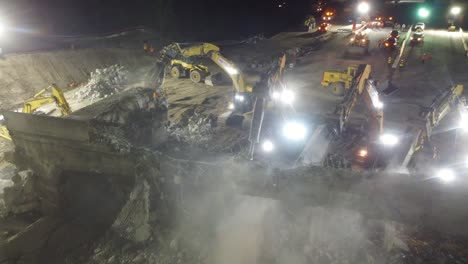 large-group-of-heavy-equipment-demolishes-a-heavily-reinforced-concrete-overpass-in-the-night,-overhead-drone-showcases-dusty-night-environment