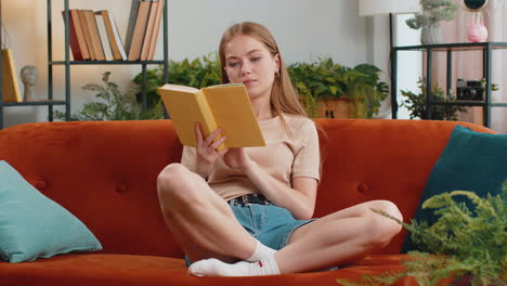 Junge-Frau-Liest-Ein-Interessantes-Buch,-Blättert-Lächelnd-Um-Und-Genießt-Die-Literatur,-Während-Sie-Sich-Auf-Der-Couch-Ausruht