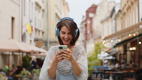 Happy-young-woman-in-wireless-headphones-choosing,-listening-music-dancing-outdoors-city-street