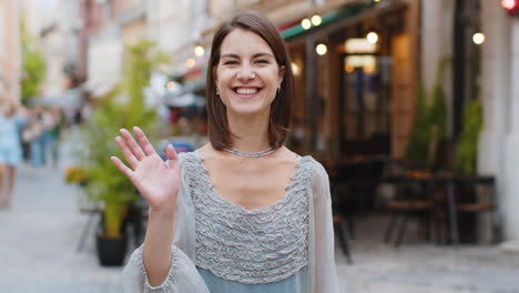 Young-woman-smiling-friendly-at-camera,-waving-hands-hello,-hi,-greeting-or-goodbye-in-city-street